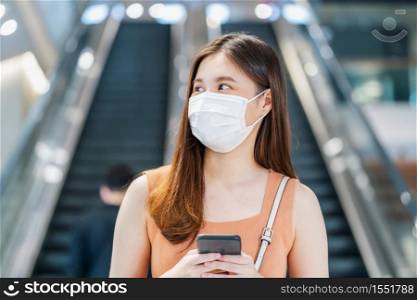 Young Asian woman passenger wearing surgical mask and using smart mobile phone in front of escalator between walk though subway interchange when traveling in big city at Covid19 outbreak, new normal