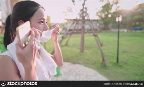 Young asian woman put on protective face mask after done exercising, sunset light on background, holding mobile phone, stay healthy during pandemic covid19 corona virus, social distancing, side view