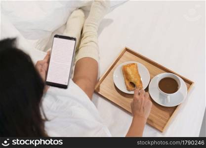 Young Asian woman relax on bed while using mobile phone and eat breakfast. Technology, lifestyle, food and beverage concept. Top view and copy space.