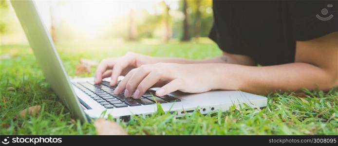 Young asian woman&rsquo;s legs on the green grass with open laptop. Girl&rsquo;s hands on keyboard. Distance learning concept. Happy hipster young asian woman working on laptop in park. Panoramic banner.