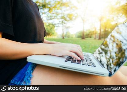 Young asian woman&rsquo;s legs on the green grass with open laptop. Girl&rsquo;s hands on keyboard. Distance learning concept. Happy hipster young asian woman working on laptop in park. Student studying outdoors.