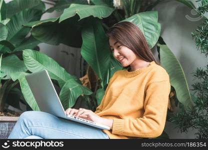 Young Asian woman shopping online with her laptop in a coffee shop garden with a background of a green tree. Young woman shopping merchandise online in the garden. Merchandise shopping online concept.