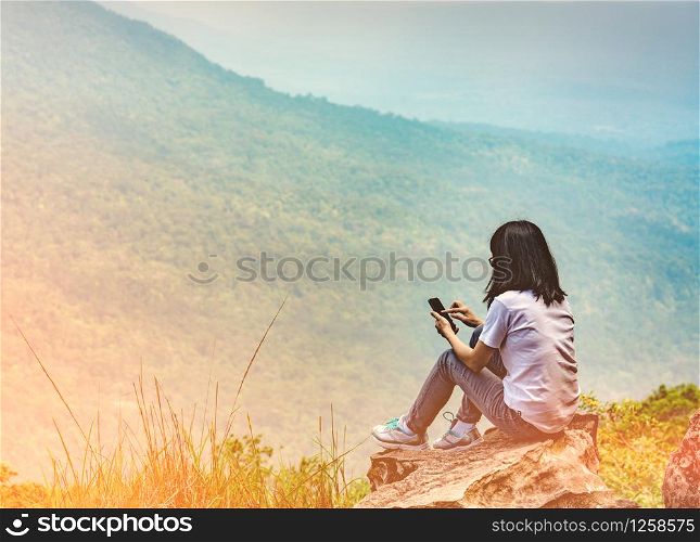 Young Asian woman sit on the rock at the cliff and using smart phone message to her friends while on vacation at the national park. Adventure travel and communication concept