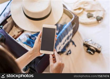 Young asian woman sitting on the bed using smartphone and packing her suitcase preparing for travel on summer vacation