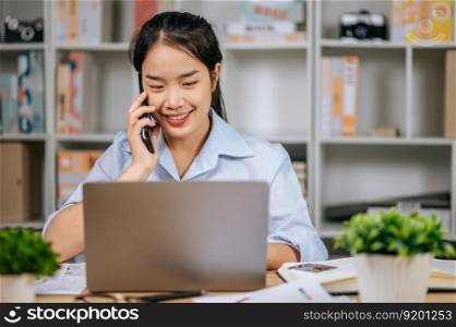 Young Asian woman smile while talking on smartphone, She working with laptop computer on desk at home office, during quarantine covid-19 self isolation at home, work from home concept