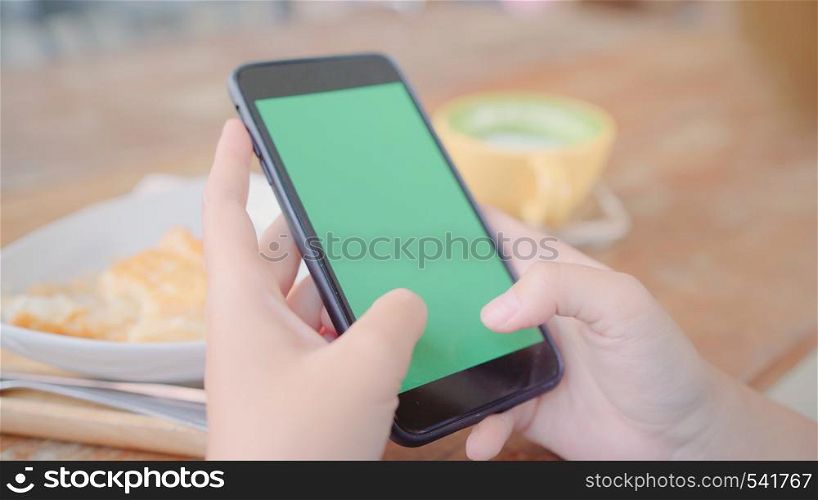 Young asian woman using black mobile phone device with green screen. Asian woman holding smartphone, scrolling pages while sitting in cafe. Chroma key.