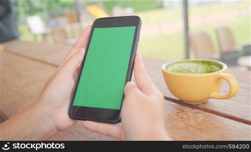 Young asian woman using black mobile phone device with green screen. Asian woman holding smartphone, scrolling pages while sitting in cafe. Chroma key.