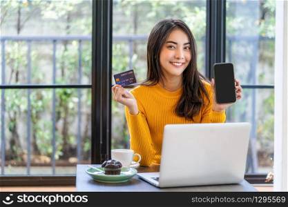 Young Asian woman using credit card and presenting mobile phone for online shopping in coffee shop or coworking space, coffee cup, technology money wallet and online payment concept, credit card mockup