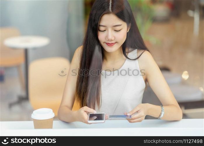 Young Asian woman using credit card with mobile phone for online shopping in coffee shop or coworking space beside window mirror, technology money wallet and online payment concept, credit card mockup