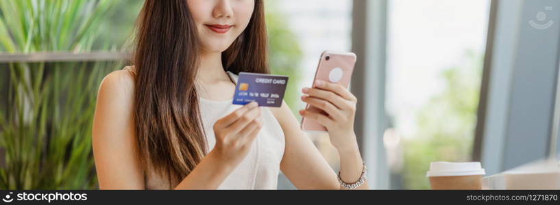 Young Asian woman using credit card with mobile phone for online shopping in coffee shop or coworking space beside window mirror, technology money wallet and online payment concept, credit card mockup