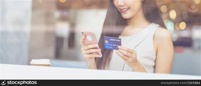 Young Asian woman using credit card with mobile phone for online shopping in coffee shop or coworking space beside window mirror, technology money wallet and online payment concept, credit card mockup
