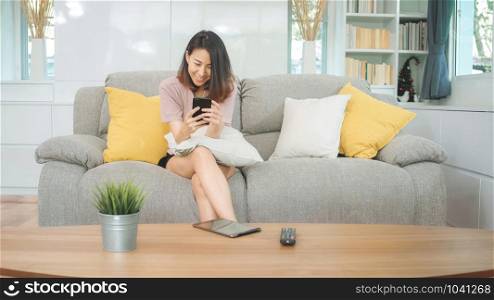 Young Asian woman using smartphone checking social media feeling happy smiling while lying on the sofa when relax in living room at home. Lifestyle latin and hispanic ethnicity women at house concept.