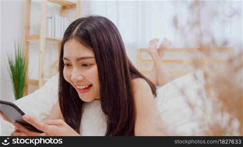 Young Asian woman using smartphone checking social media feeling happy smiling while lying on bed after wake up in the morning, Beautiful attractive Japanese girl smiling relax in bedroom at home.