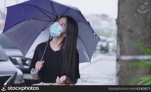 young asian Woman wear protective face mask holding blue umbrella standing on the street side walk, shower rainy season, pouring rain, risk of getting sick, hard pouring rain, reach out hand curious