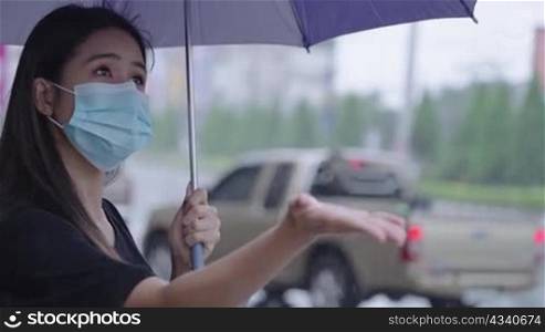 young asian Woman wear surgical mask holding umbrella standing on the road side, on the raining day, shower rainy season tropical weather, reach out her hand feeling the rain drops, protect her self