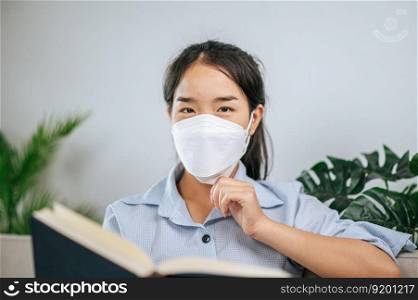 Young asian woman wearing face mask sitting on sofa in the living room, she reading book during quaranti≠covid-19 self isolation at home