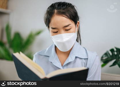 Young asian woman wearing face mask sitting on sofa in the living room, she reading book during quarantine covid-19 self isolation at home
