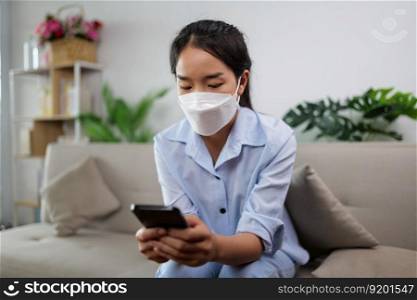 Young asian woman wearing face mask sitting on sofa in the living room, she use smartphone chatting during quarantine covid-19 self isolation at home