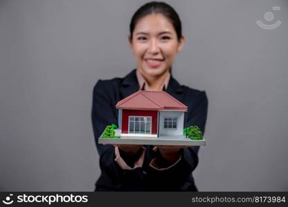 Young Asian woman wearing formal suit, holding and showcasing house model on isolated background. Ideal for housing advertisement, home insurance promotions, or customizable copy space. Enthusiastic. Young Asian woman wearing formal suit holding house model. Enthusiastic