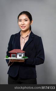 Young Asian woman wearing formal suit, holding and showcasing house model on isolated background. Ideal for housing advertisement, home insurance promotions, or customizable copy space. Enthusiastic. Young Asian woman wearing formal suit holding house model. Enthusiastic