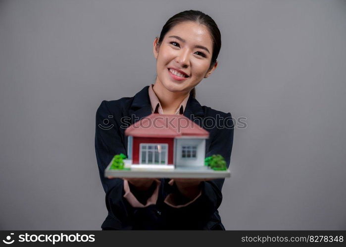 Young Asian woman wearing formal suit, holding and showcasing house model on isolated background. Ideal for housing advertisement, home insurance promotions, or customizable copy space. Enthusiastic. Young Asian woman wearing formal suit holding house model. Enthusiastic