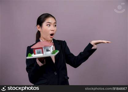 Young Asian woman wearing formal suit, holding and showcasing house model on isolated background. Ideal for housing advertisement, home insurance promotions, or customizable copy space. Enthusiastic. Young Asian woman wearing formal suit holding house model. Enthusiastic