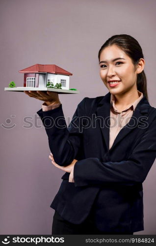 Young Asian woman wearing formal suit, holding and showcasing house model on isolated background. Ideal for housing advertisement, home insurance promotions, or customizable copy space. Enthusiastic. Young Asian woman wearing formal suit holding house model. Enthusiastic