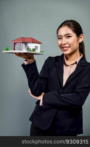 Young Asian woman wearing formal suit, holding and showcasing house model on isolated background. Ideal for housing advertisement, home insurance promotions, or customizable copy space. Enthusiastic. Young Asian woman wearing formal suit holding house model. Enthusiastic