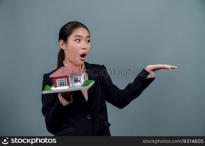 Young Asian woman wearing formal suit, holding and showcasing house model on isolated background. Ideal for housing advertisement, home insurance promotions, or customizable copy space. Enthusiastic. Young Asian woman wearing formal suit holding house model. Enthusiastic