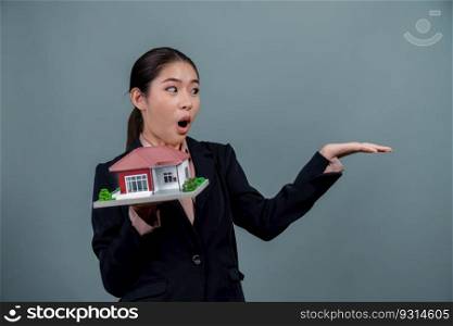 Young Asian woman wearing formal suit, holding and showcasing house model on isolated background. Ideal for housing advertisement, home insurance promotions, or customizable copy space. Enthusiastic. Young Asian woman wearing formal suit holding house model. Enthusiastic