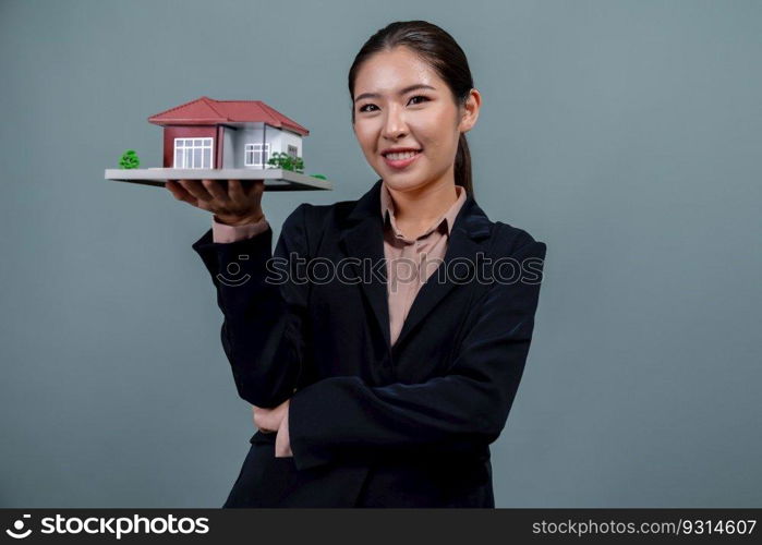 Young Asian woman wearing formal suit, holding and showcasing house model on isolated background. Ideal for housing advertisement, home insurance promotions, or customizable copy space. Enthusiastic. Young Asian woman wearing formal suit holding house model. Enthusiastic