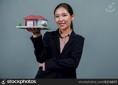 Young Asian woman wearing formal suit, holding and showcasing house model on isolated background. Ideal for housing advertisement, home insurance promotions, or customizable copy space. Enthusiastic. Young Asian woman wearing formal suit holding house model. Enthusiastic