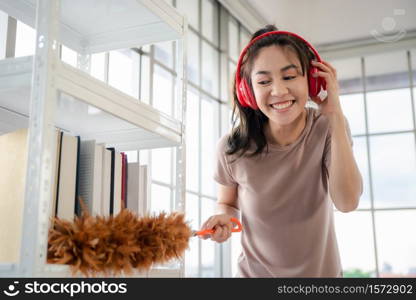 Young asian woman with headphones using a feather duster to clean wood book shelves in home .