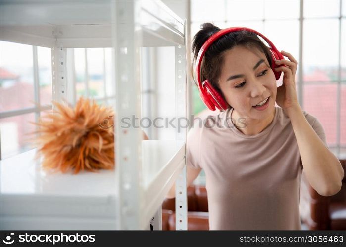 Young asian woman with headphones using a feather duster to clean wood book shelves in home .