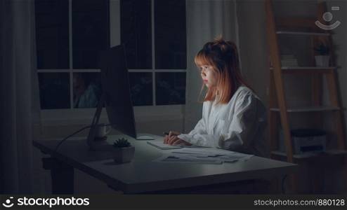 Young asian woman working late using desktop on desk in living room at home. Asia business woman writing notebook document finance and calculator in night at home office. Overworked female concept.
