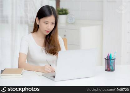 Young asian woman writing on notebook for planning working and using laptop computer on desk at home, girl notes about finance, female study and learning, business and communication concept.