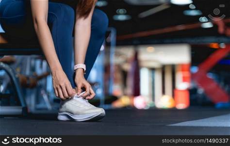 Young Asian Women are tying shoelaces To prepare before exercise and for safety, A healthy woman sitting on the bench leaning down to tie the shoelace at gym with blurback ground and copy space