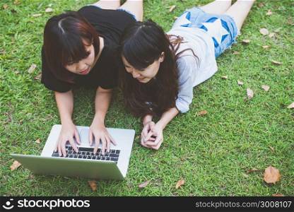 Young asian women lying on grass and using laptop and typing. Girls hands on keyboard. Distance learning concept. Happy hipster young asian women working on laptop in park. Student studying outdoors.