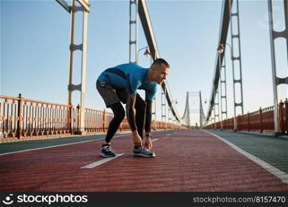 Young athlete man tying lace on running shoes ready for morning jog. Sportsman runner training on urban bridge. Young athlete man tying lace on running shoes during training on city bridge