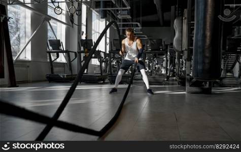Young athletic man using ropes to build arm strength doing cross fit training exercise in gym. Strength and motivation concept. Athletic man using ropes doing cross training exercise in modern gym
