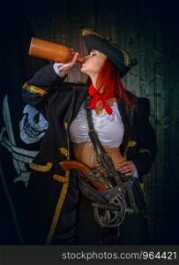 Young Attractive Armed Girl Pirate Captain Drinks Clay Bottle Alcohol Jolly Roger Flag Background. Girl Pirate Captain