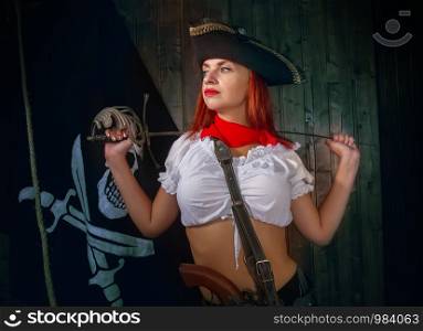 Young attractive armed girl pirate captain looks into the distance on the background of the flag Jolly Roger. Girl Pirate Captain