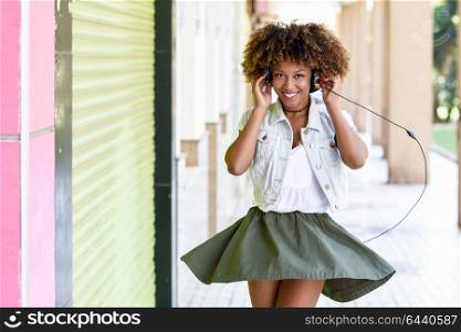 Young attractive black woman in urban street listening to the music with headphones. Girl wearing casual clothes with afro hairstyle