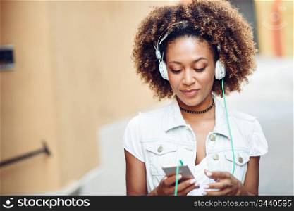 Young attractive black woman in urban street listening to the music with headphones and smartphone. Girl wearing casual clothes with afro hairstyle