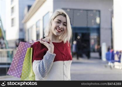 Young attractive blonde woman in a read and white sweater against the backdrop of the city streets. Holding a lot of shopping bags.