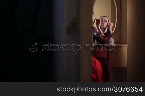 Young attractive blonde woman sitting at the dressing table with Christmas decorations, different brushes and applying eyebrow makeup. Sexy lady defining and shaping her eyebrows with a brush looking at the mirror.