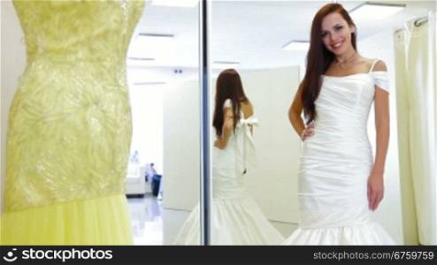 Young attractive bride trying on wedding gown in bridal shop, posing and looking at camera