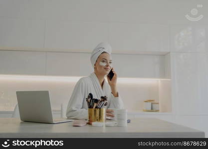 Young attractive business woman in bathrobe and towel turban on head with cosmetic patches under eyes talking on mobile phone and working on laptop while getting ready for work in morning at home. Beautiful woman in bathrobe with towel on head talking on mobile phone while doing skin care procedures at home