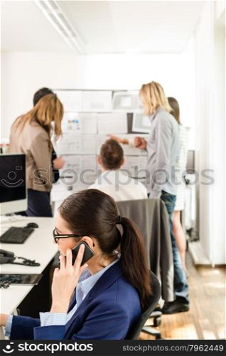 Young attractive business woman sitting in front of her computer with her mobile phone to her ear in stylish office with group discussing conceptual papers in the background, might be a startup or a creative agency