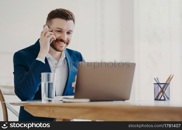 Young attractive businessman in formal suit smiling when talking on phone and looking at laptop screen approving what he sees, ceo sitting in minimalistic office environment with stylish work place. Young stylish executive talking on phone in front of laptop behind office desk